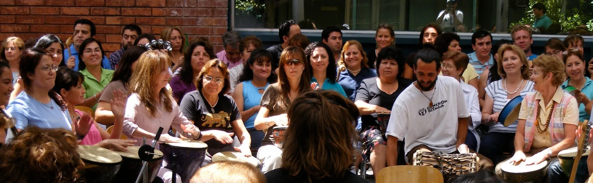 Un círculo grande de personas sonriendo mientras tocan tambores al aire libre, frente a un muro de ladrillo; un facilitador con la polera de Conexión Rítmica toca un tambor en primer plano.