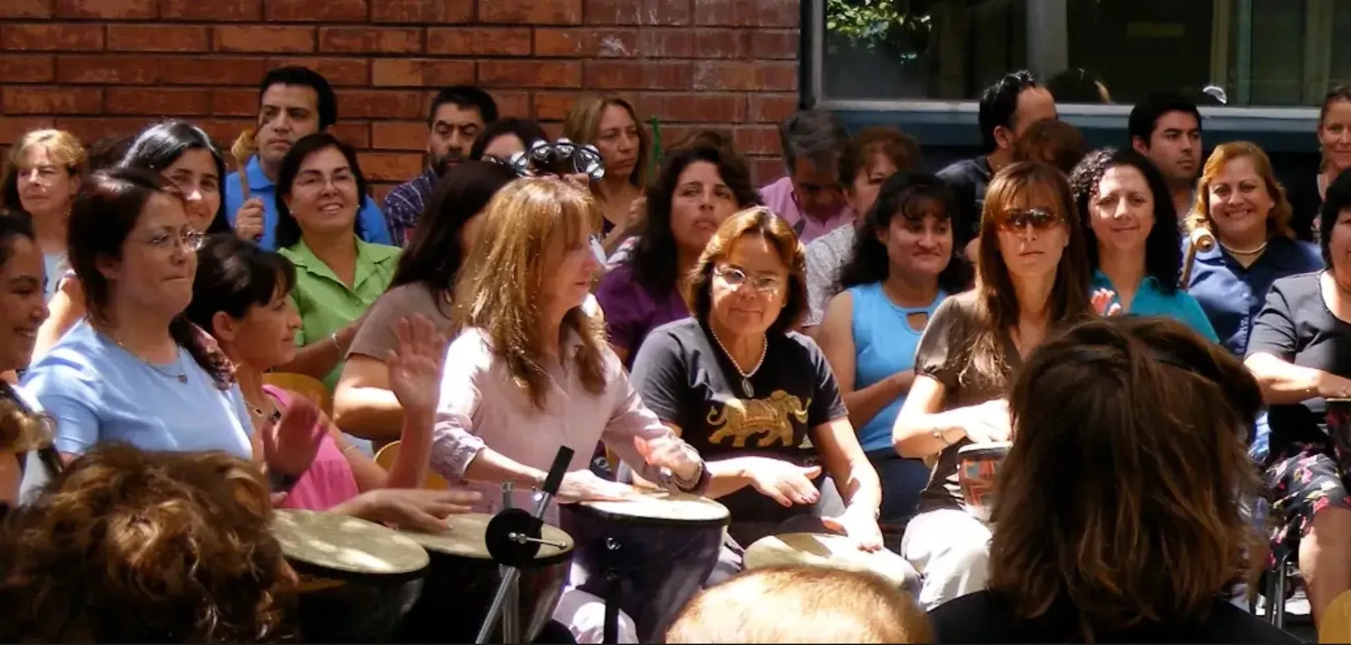 Un grupo numeroso de personas sonrientes sentadas en círculo al aire libre frente a un muro de ladrillo, tocando tambores y percusión.